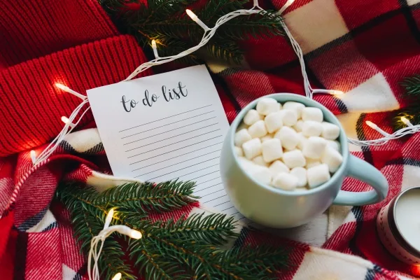 Holiday to do list next to a cup of hot chocolate with marshmallows and Christmas decoration on the red plaid background.