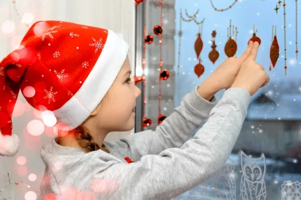 Little girl in a Santa hat rejoices as she sticks a Christmas snowflake sticker on her window at home.